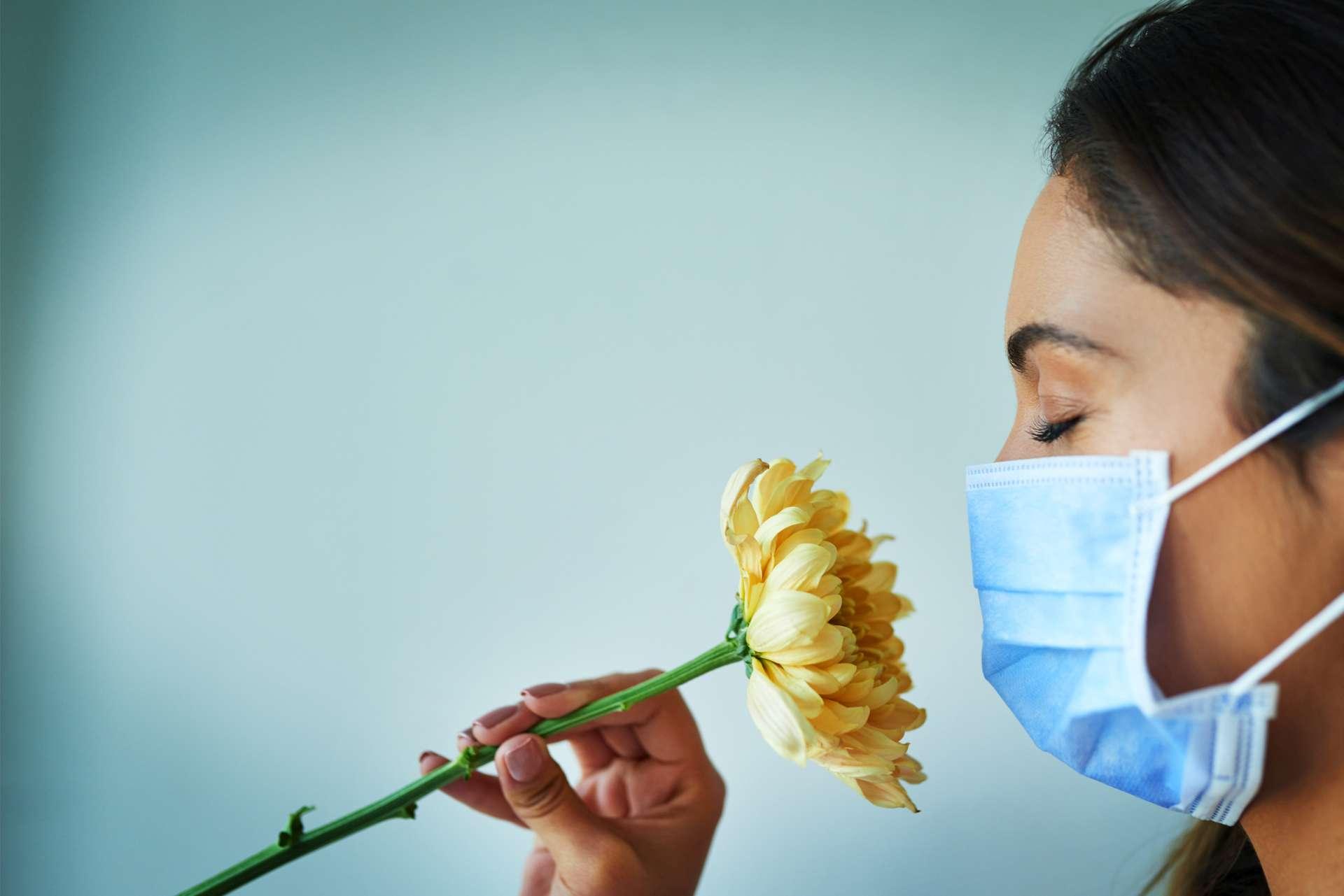 Schuss einer jungen Frau, die eine Blume riecht, während sie eine chirurgische Maske trägt