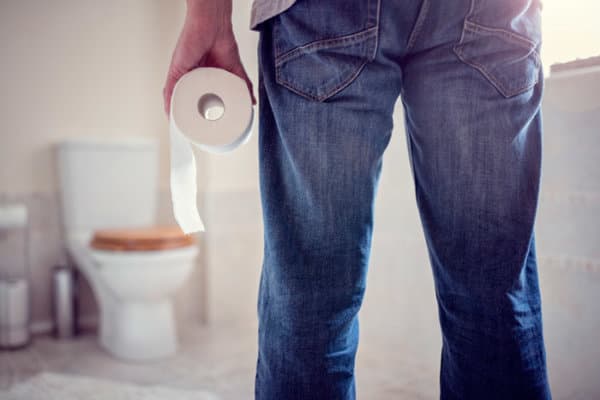 Mann hält Toilettenpapierrolle im Badezimmer und schaut auf die Toilette